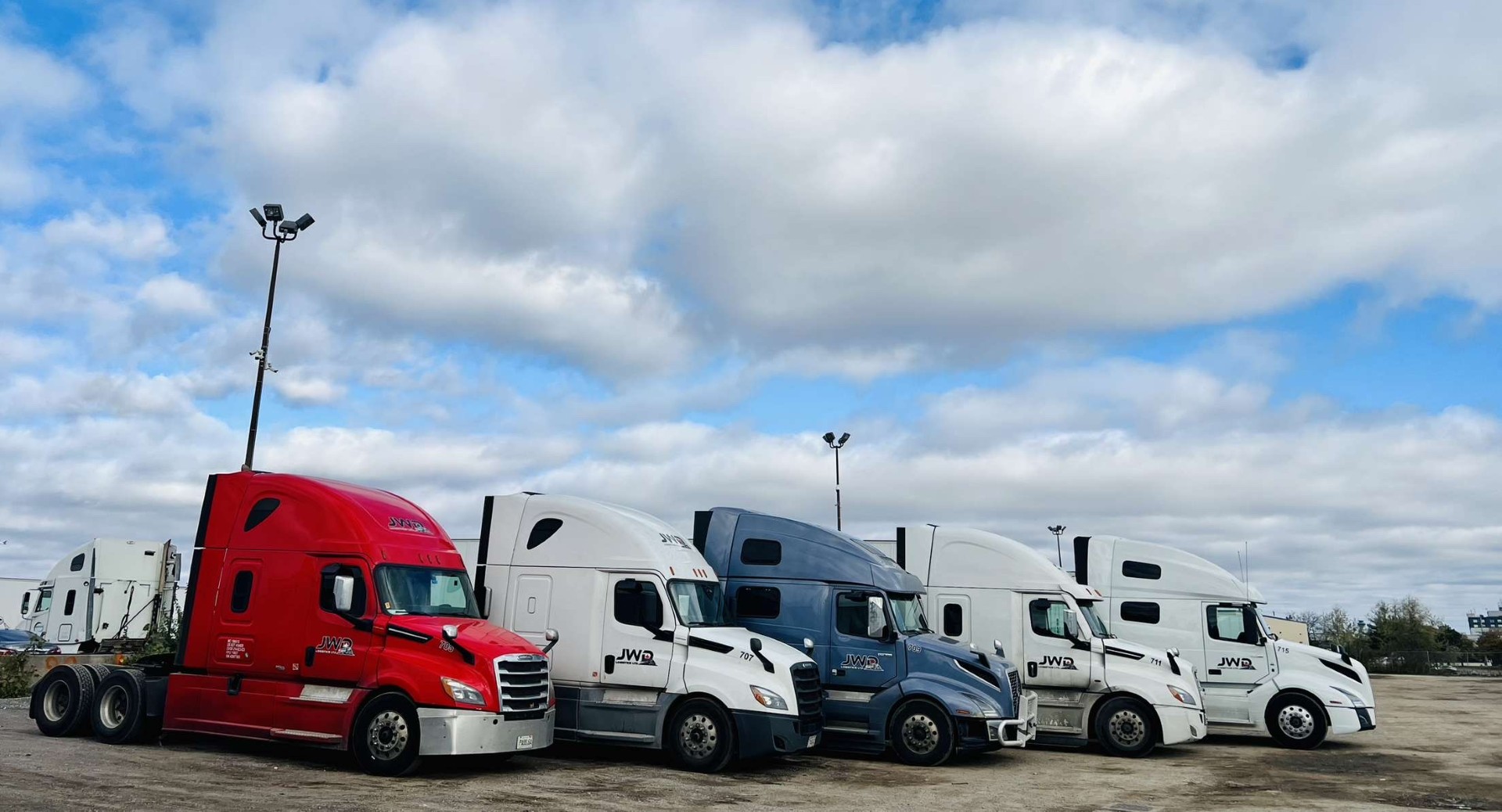 Five JWD Logistics tractors in a row — red, white, navy, and white cabs — parked at the terminal yard.