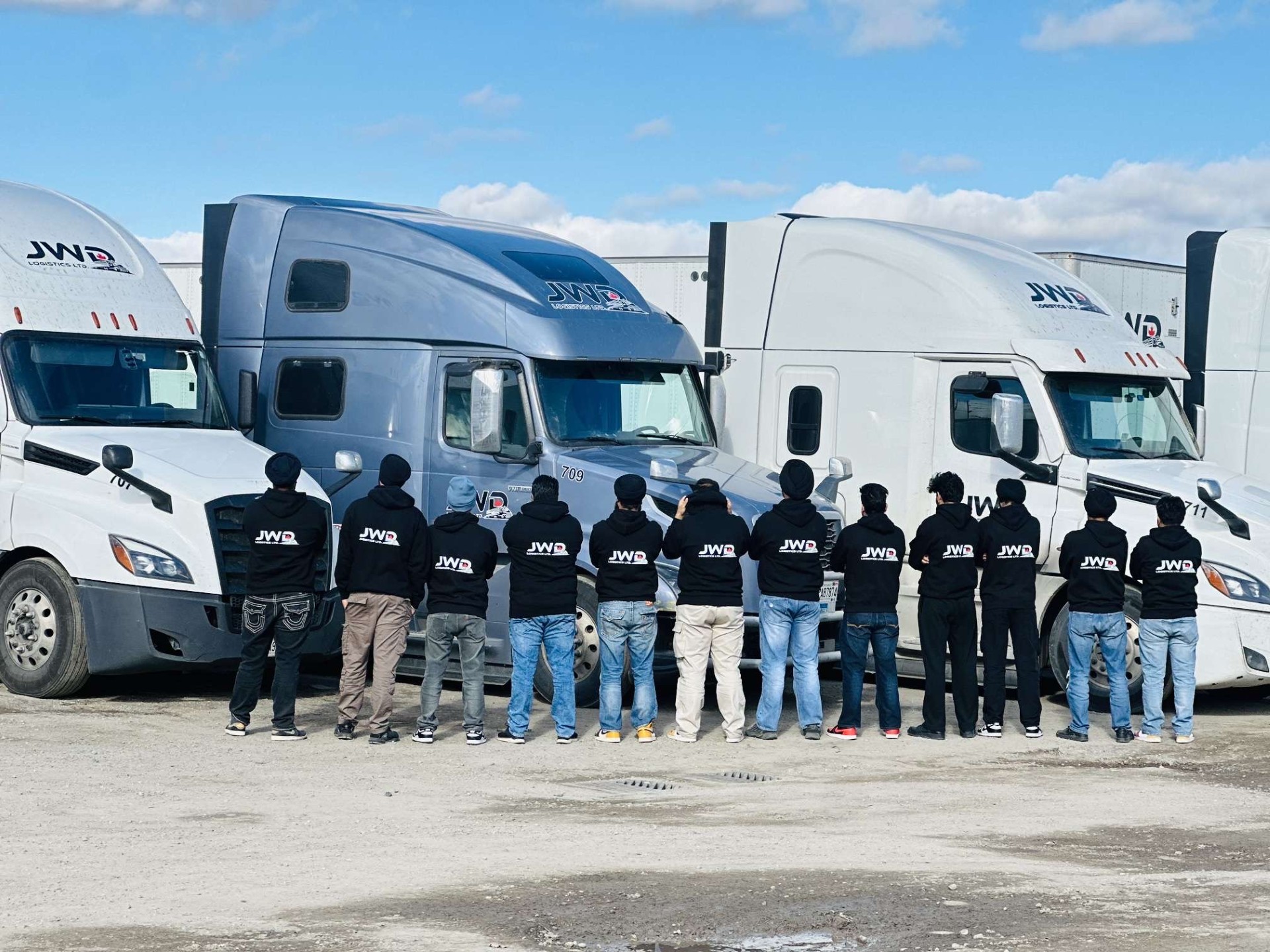 Eleven JWD Logistics drivers in matching JWD hoodies standing in front of the fleet at the terminal yard.